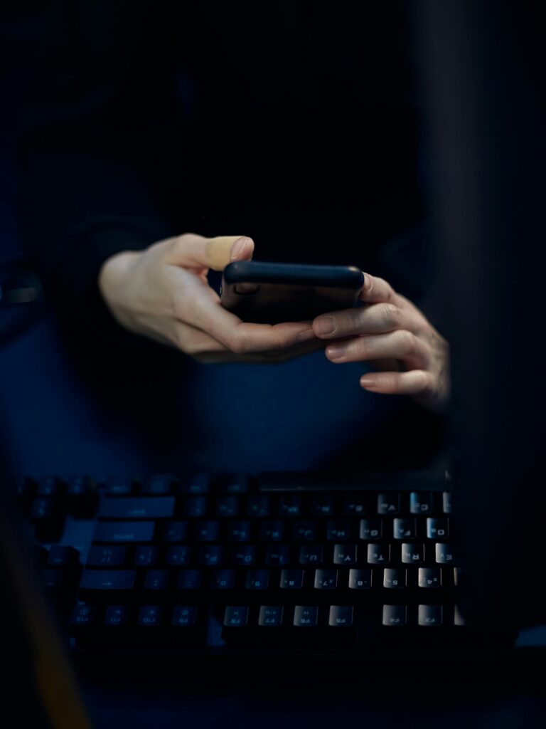 Close-up of hands using a smartphone in a dimly lit room with a computer keyboard.